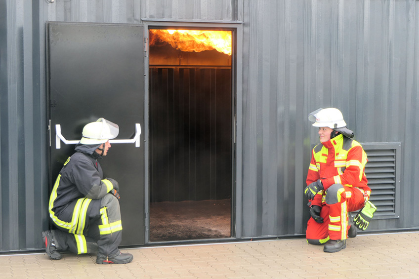 Aus einem Brandsimulationscontainer schießt Feuer von der Decke innen nach außen. Vor der Tür hocken zwei Brandmeisteranwärter aus Minden und Porta Westfalica in Schutzkleidung und warten darauf, in die „heiße Hölle“ zu stürmen.
