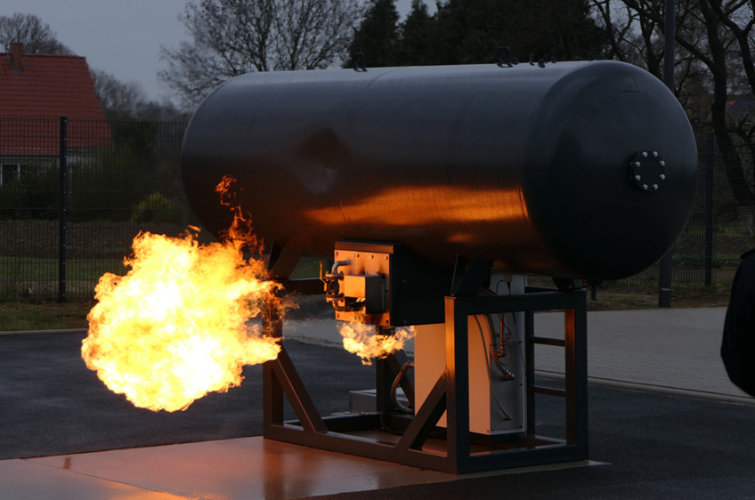 Aus einem Brandsimulationscontainer schießt Feuer von der Decke innen nach außen. Vor der Tür hocken zwei Brandmeisteranwärter aus Minden und Porta Westfalica in Schutzkleidung und warten darauf, in die „heiße Hölle“ zu stürmen.