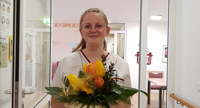  Liane Schellenberg ist Auszubildende zur Pflegefachkraft im Volkeninghaus, einer Senioreneinrichtung des Ludwig-Steil-Hofs. Sie &uuml;berreicht einen Blumenstrau&szlig; und Gr&uuml;&szlig;e an einen Bewohner, dessen Angeh&ouml;rige leider nicht pers&ouml;nlich kommen konnten.